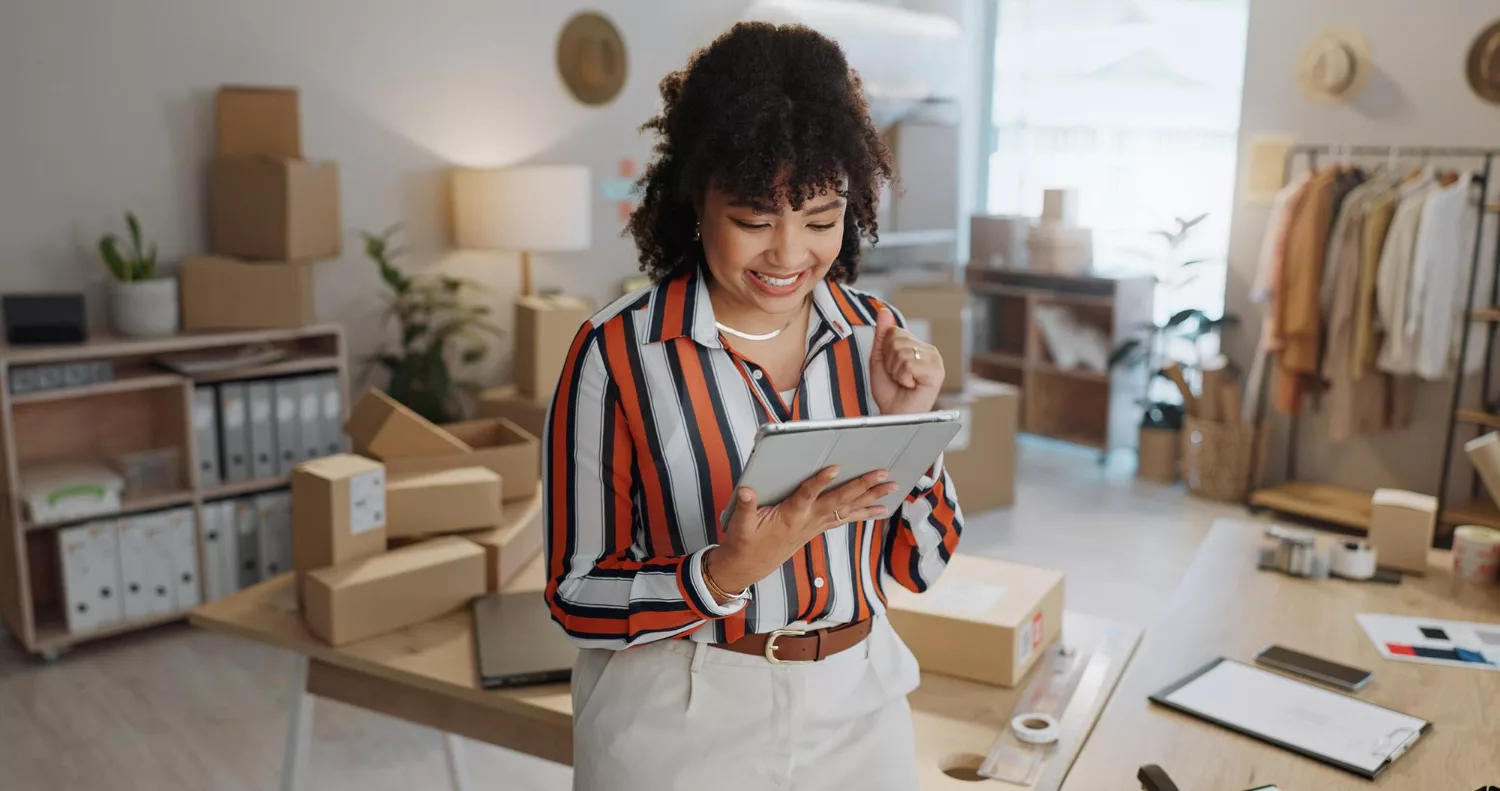 Small business owner using a tablet in an office surrounded by shipping boxes while managing AI automation workflows.